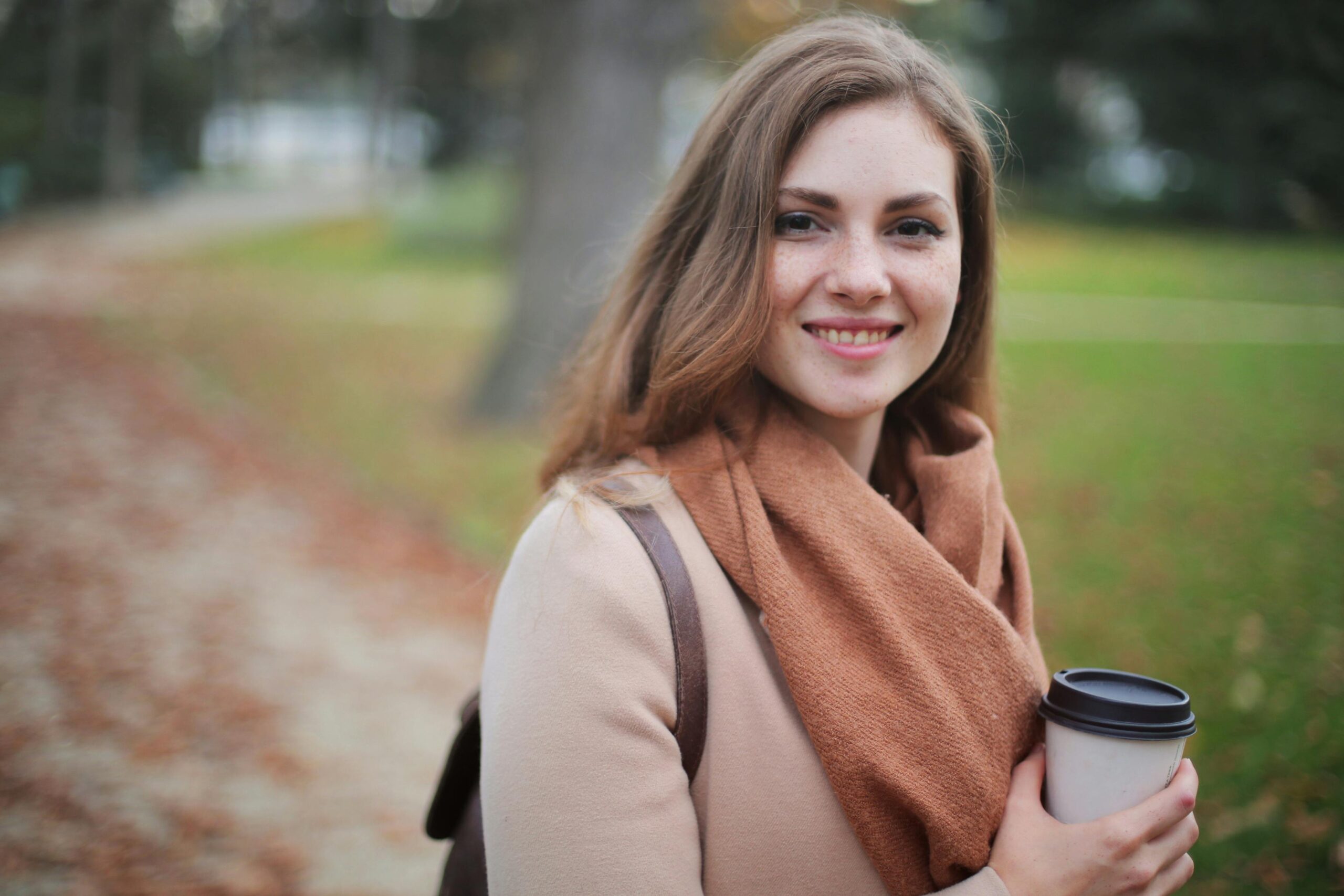 Portrait of a smiling woman holding a coffee cup in a serene autumn park setting.